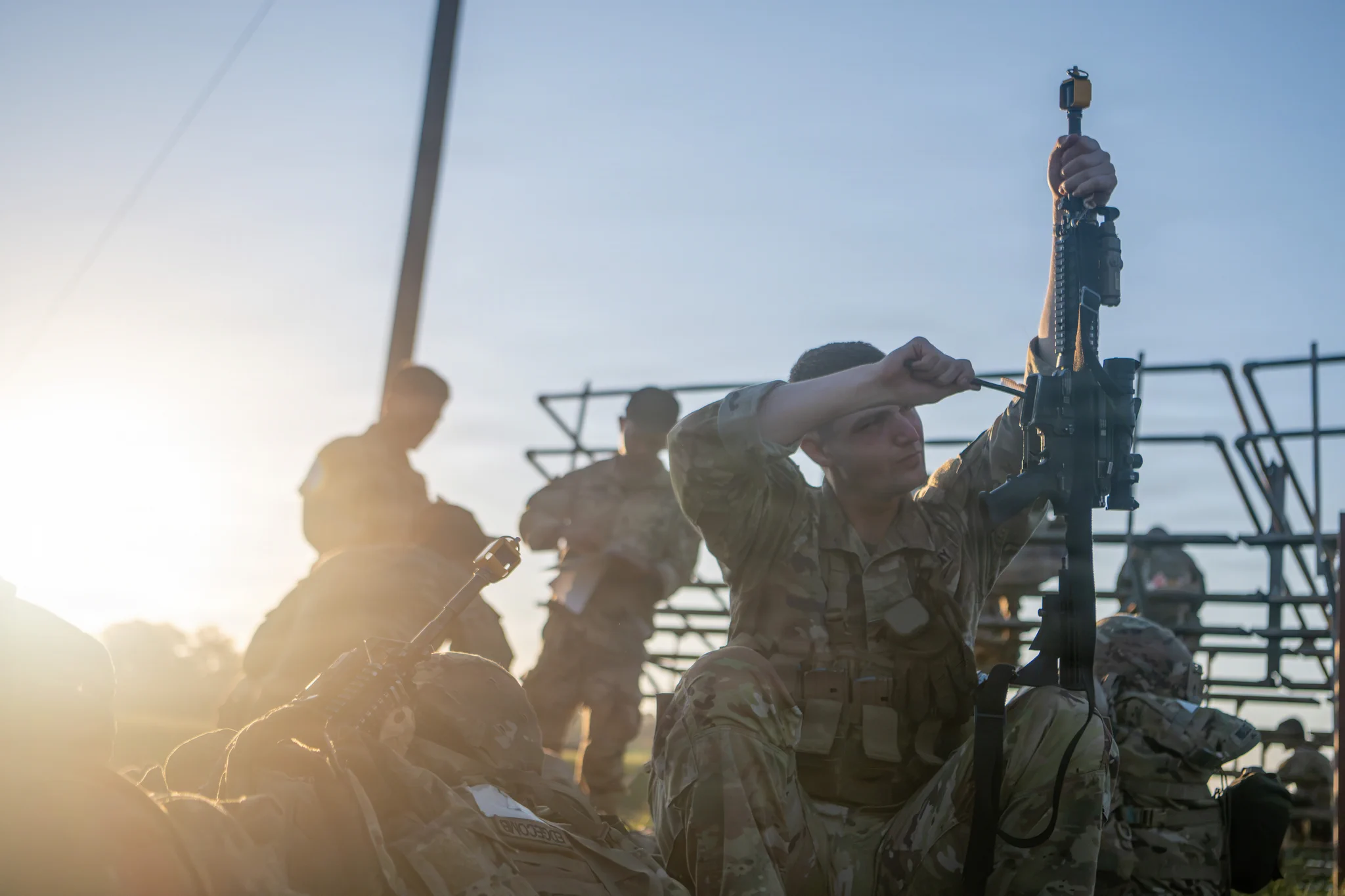 A male soldier in camouflage kneels, intently cleaning his rifle with a brush as sunlight flares from the left. His face is visible, focused on the weapon. Other blurry soldiers and training structures are in the background. 
