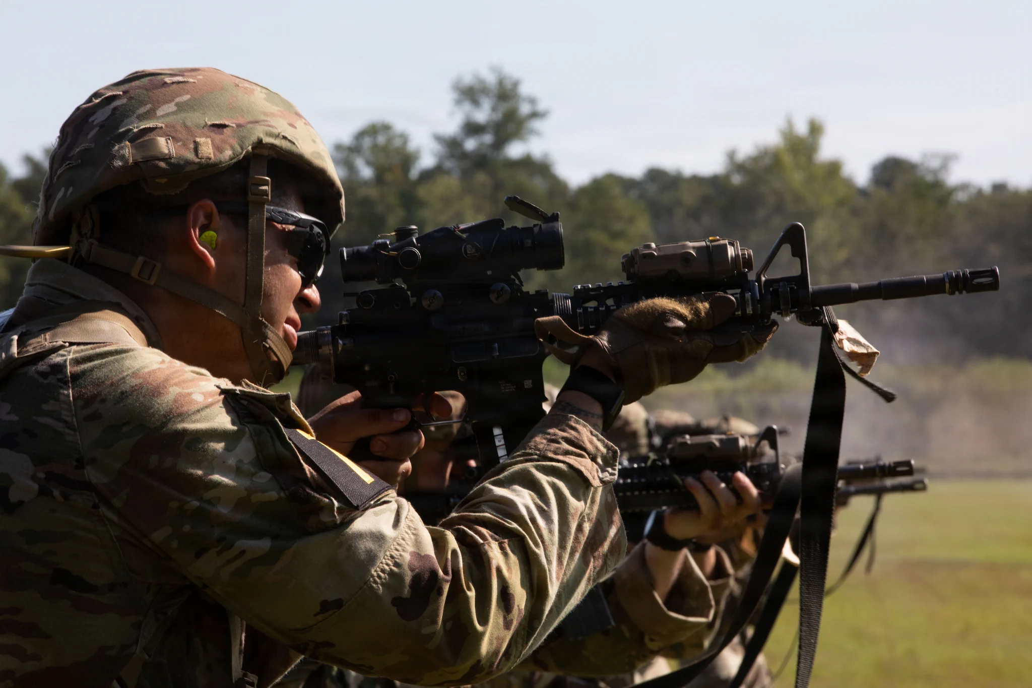 A soldier in camouflage uniform, helmet, safety glasses, and yellow earplugs aims an assault rifle. Seen from the side, they hold the rifle with both hands, aiming forward. Behind them, other soldiers also aim, with dust rising from the firing line on a sunny day in a grassy field with trees.