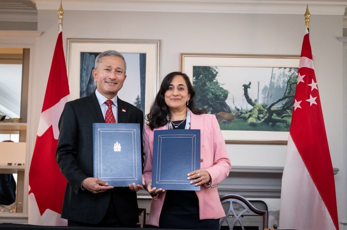 La ministre Anand et le ministre Balakrishnan tenant des dossiers devant les drapeaux canadien et singapourien.