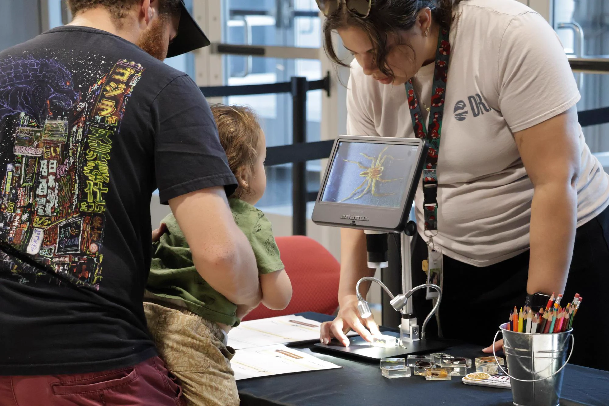 A person is holding a child near a table where another person is placing a slide under a microscope and the screen displays an image of an insect.