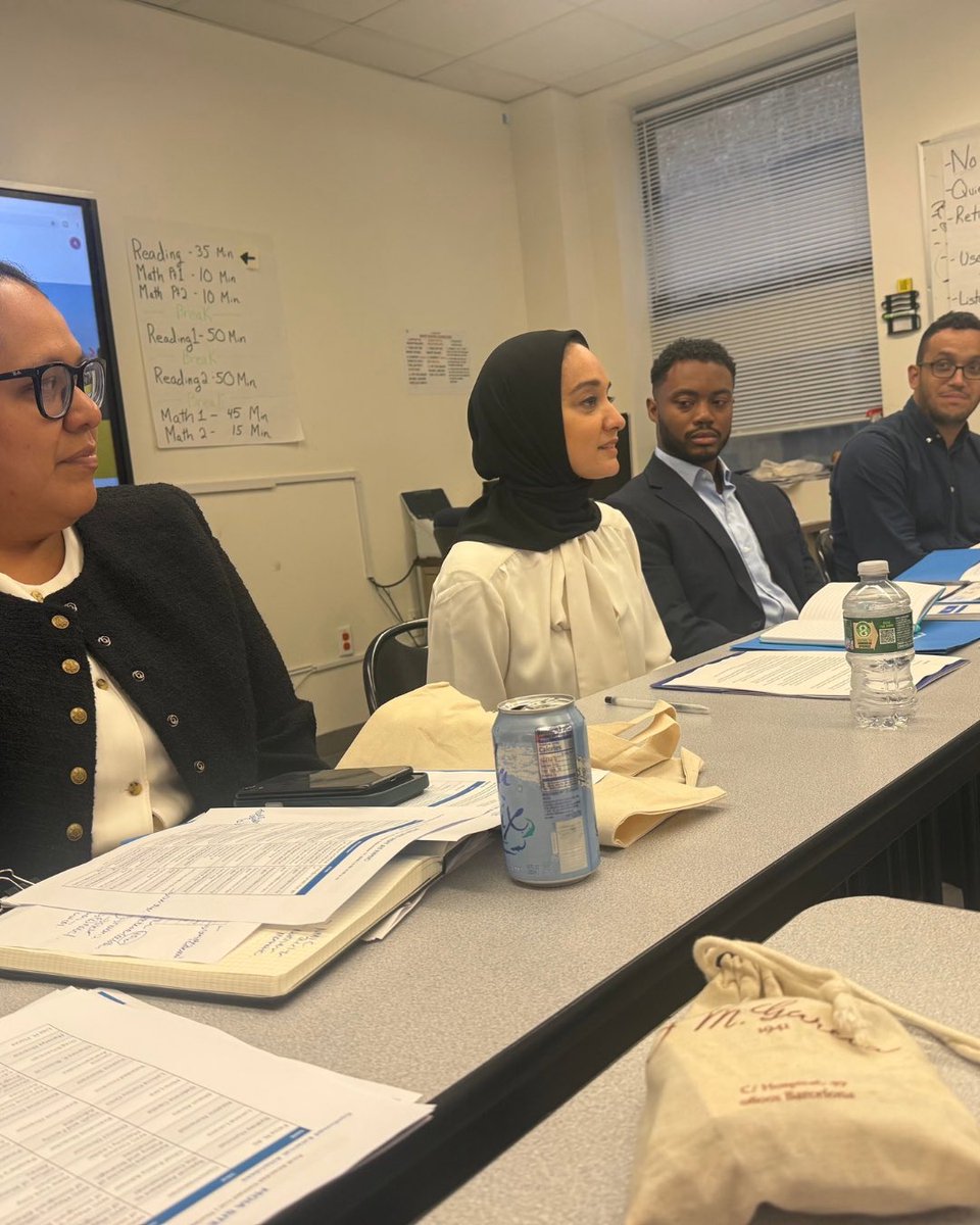 Several people sit around a long table in a meeting room. Papers, drinks, and folders are placed on the table in front of them. There is a whiteboard and window in the background.