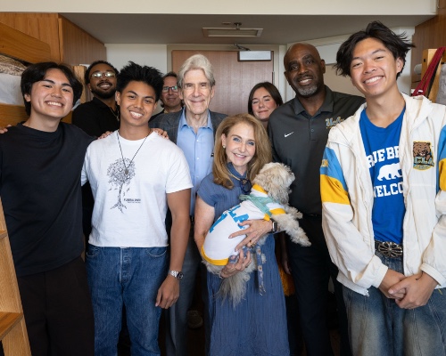 A group of students and adults pose together inside a residence hall dorm room with wooden bunk beds. A woman in front holds a small dog wearing a UCLA shirt.