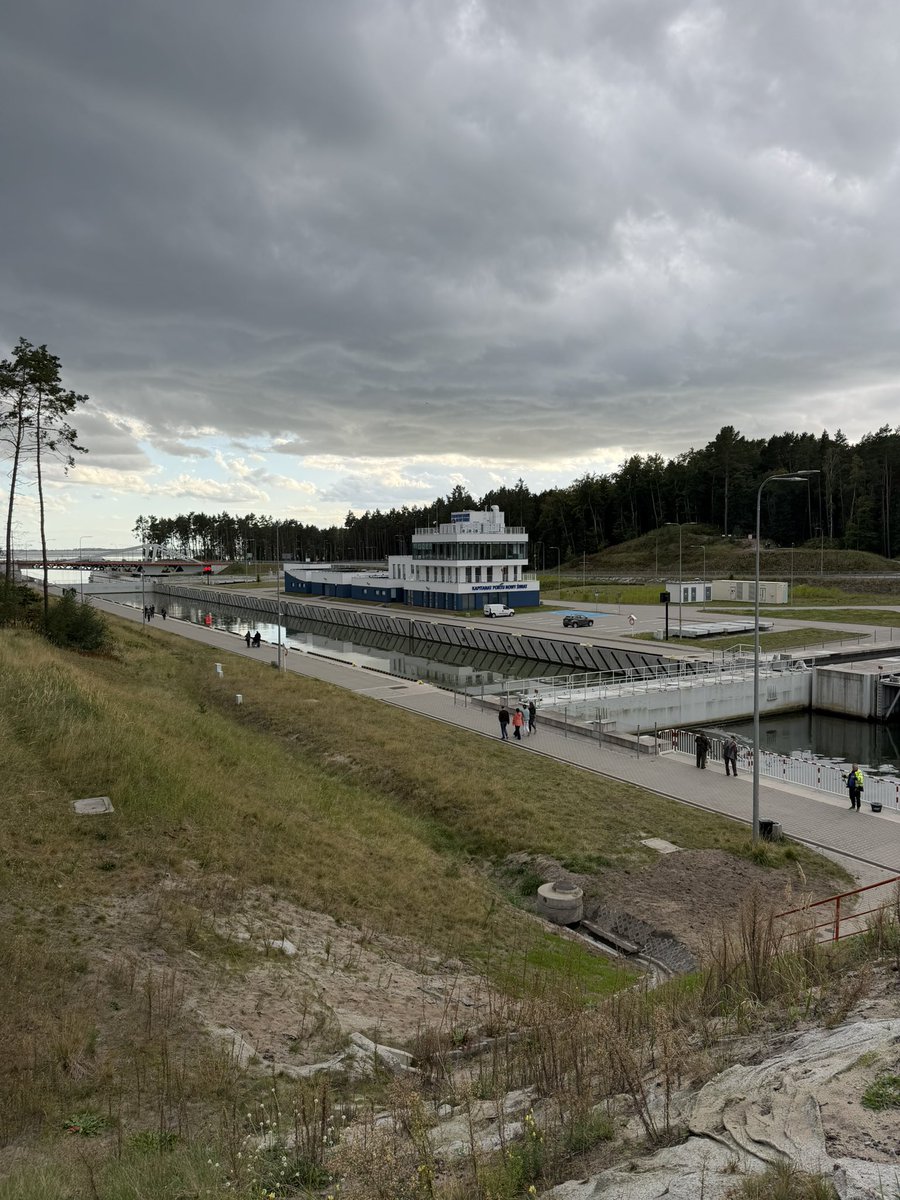 ZawadkaMarek's tweet photo. Przekop Mierzei Wiślanej

Nowe to i ładne. W końcu zrealizowana inwestycja hydrotechniczna. Szybko i sprawnie, jak na budowlę hydro.

Nowocześnie rozplanowane drogi, ścieżki do dreptania i jazdy rowerem. Generalnie fajnie pod turystów i po to, żeby pokazać, że sensownie wydano publiczne pieniądze.

Można oczywiście mieć z tyłu głowy wątpliwości. Choćby tę: czy zamiast tego nie należało zainwestować w Zalew Szczeciński i budować tam realnej alternatywy dla Trójmiasta. Tym bardziej że od 1946 r. nikt w Polsce nie poddał całościowej rewizji zagospodarowania naszej przestrzeni portowej.

Ale fakt jest faktem: przekop istnieje. Jest nowy i ładny, a budynek kapitanatu zaskakująco udany architektonicznie.

I tak, spacerując po tym wszystkim w ramach konferencji, włączył mi się tryb: benchmarking muzealny.

Oczywiście zasięg GSM ledwo zipie. Po co komu zasięg? Choćby po to, żeby stojąc na wietrze, dowiedzieć się czegoś o konstrukcji, którą właśnie ogląda. Bo informacji na terenie przekopu jakby nie ma. 

Tablice informacyjne? Owszem, są. Dwie.

Zdjęcie i sporo cyfr. Inżynier podał parametry, grafik wrzucił je na szablon, dodano fotografię i wymóg informacyjny inwestycji został spełniony. Informacja odhaczona.

Czy można inaczej? Oczywiście.

W Rotterdamie zapora Maeslantkering ma kapitalną strefę informacyjno-dydaktyczną oraz plenerową ścieżkę pokazującą obieg wody w przyrodzie. Za daleko? To spójrzmy na podnośnię statków Niederfinow.

Skoro u nas nie ma dedykowanego centrum edukacyjnego i zapewne długo nie będzie, co można zrobić po kosztach?

Postawić kilkanaście spójnych tablic opowiadających o tym, czym ten przekop właściwie jest. Powiedzmy po cztery lub pięć po każdej stronie.

1. Od strony Elbląga

Co dalej ze szlakiem wodnym? Co z portem w Elblągu? Tu aż się prosi o pokazanie skali całego przedsięwzięcia. Do tego plansze wyjaśniające, czym są Żuławy oraz ile dekad projektowano kanał przez Mierzeję.

2. Przy samej śluzie

Cztery plansze pokazujące krok po kroku etapy budowy, schemat działania komory oraz mechanizm wrót.

Interaktywny model, którym mogłyby bawić się dzieci, byłby luksusem. Trzymajmy się jednak wariantu budżetowego.

3. Przy wyjściu na Zatokę Gdańską

Po lewej stronie: panorama Trójmiasta z opisem widocznych w dali konstrukcji i portów.

Po prawej: kierunek Królewiec oraz geopolityczny kontekst rosyjskiej bazy morskiej i toru wodnego, od którego przekop nas uniezależnił.

4. Pomiędzy nimi

Dwie plansze o przyrodniczej specyfice Mierzei oraz kilka dynamicznych kadrów ze śluzowania jednostek handlowych i technicznych, okrętów oraz jachtów.

I jeszcze detal czysto ludzki: infrastruktura.

Szukałem ławek, bo spacerowało tam wielu starszych ludzi. Nie znalazłem ani jednej poza strefą gastronomiczną. Z toaletami ten sam problem, skupiono je w jednym miejscu.

Aż się prosi o ławki wzdłuż ciągów spacerowych oraz przynajmniej jeden punkt sanitarny po każdej stronie kanału.

Przy nabrzeżu mogłaby też czasami cumować jednostka Straży Granicznej, Marynarki Wojennej albo Urzędu Morskiego, stając się żywym elementem opowieści o tym miejscu. Dla decydentów to zapewne fanaberia.

Tak bym to zrobił na miejscu gospodarzy tego terenu.

Bo skoro wydano potężne publiczne pieniądze i naprawdę jest się czym chwalić, wypadałoby wreszcie nauczyć się o tym opowiadać. I się chwalić. Jest czym. 

@marekgrobarczyk @MI_GOV_PL
