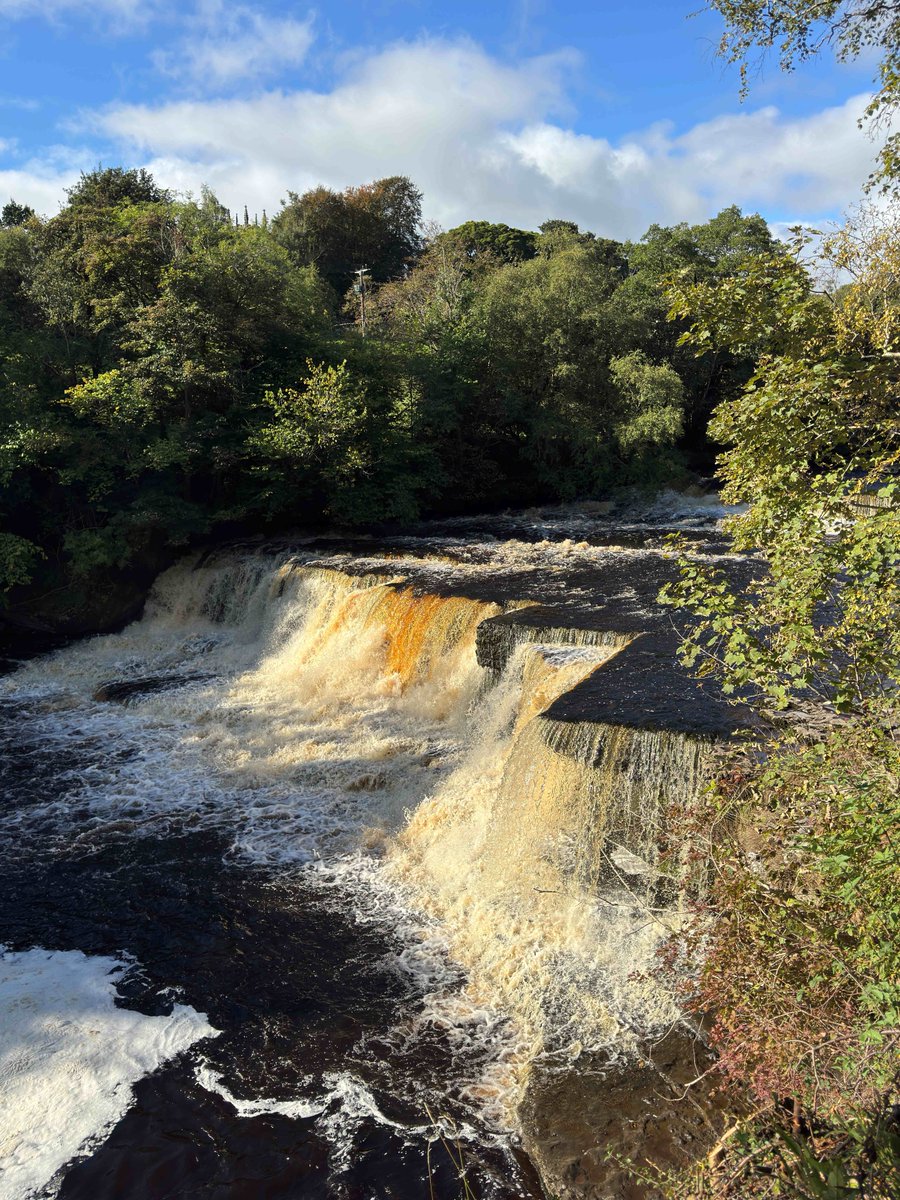 Wide waterfall cascading over layered rocks surrounded by dense green trees under a partly cloudy blue sky.