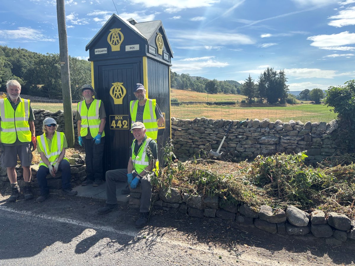 Five volunteers in yellow safety vests posing beside a vintage black AA roadside emergency box numbered 442 on a sunny rural roadside.