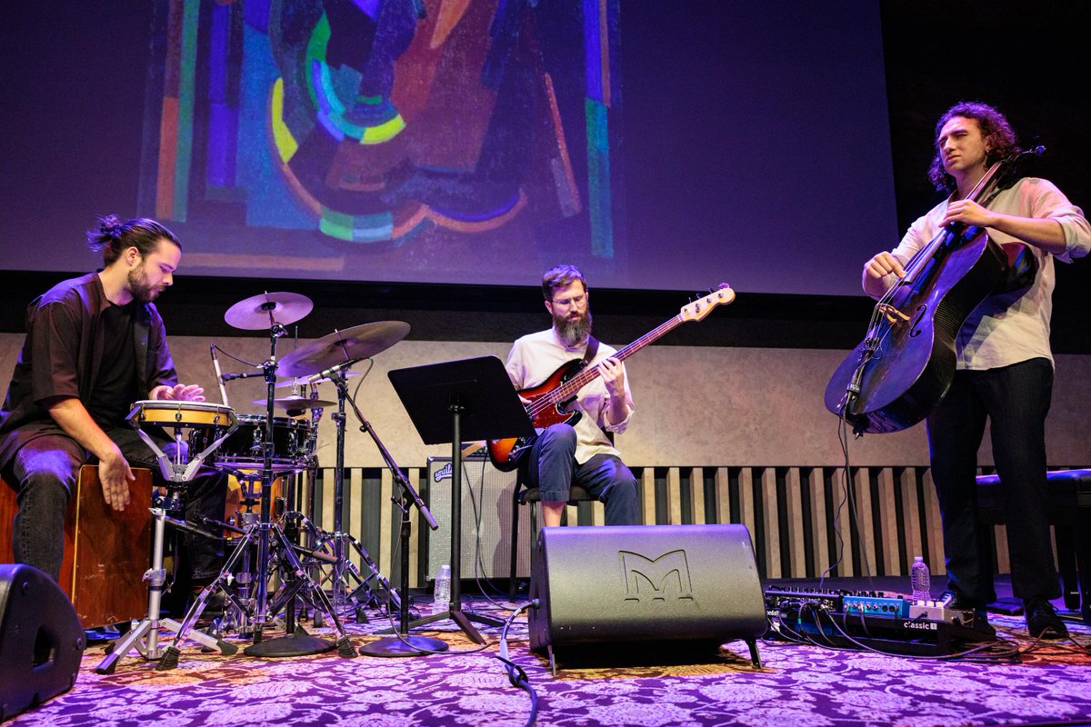 A musical trio performs on stage, featuring a percussionist, a guitarist, and a cellist. The guitarist holds an electric guitar, while the percussionist plays drums and the cellist stands with a cello. Abstract artwork is projected in the background. The setup includes microphones, amplifiers, and cables on a patterned carpet.