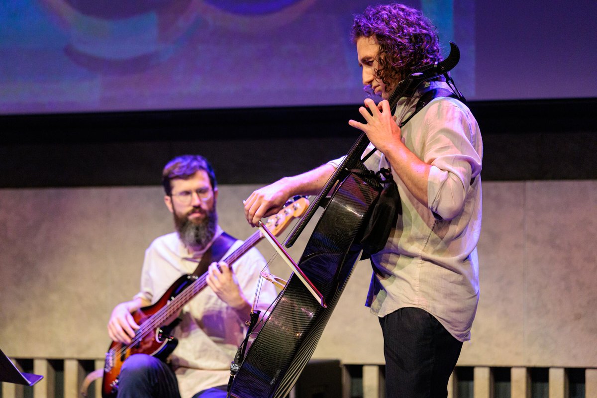 Two individuals are performing on stage with musical instruments. The person on the left is playing a bass guitar, while the person on the right is playing an electronic cello. The setting appears to be a live music event.