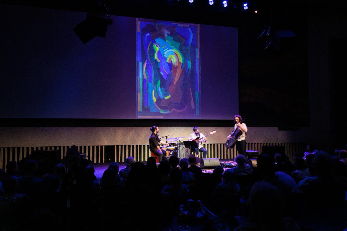 A musical trio performs on stage in a dimly lit venue, with a colorful abstract painting projected behind them. An audience is seated in the foreground, watching attentively.