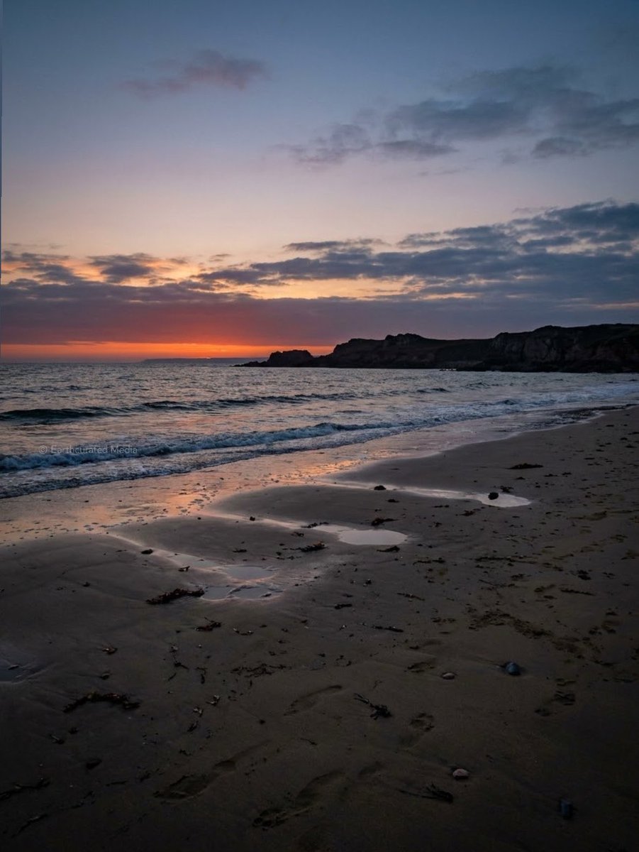 Original media by @earthcurated. Sunset over a quiet beach, with warm orange light beneath dark clouds reflecting across the wet shoreline. © Earthcurated Media. All rights reserved.