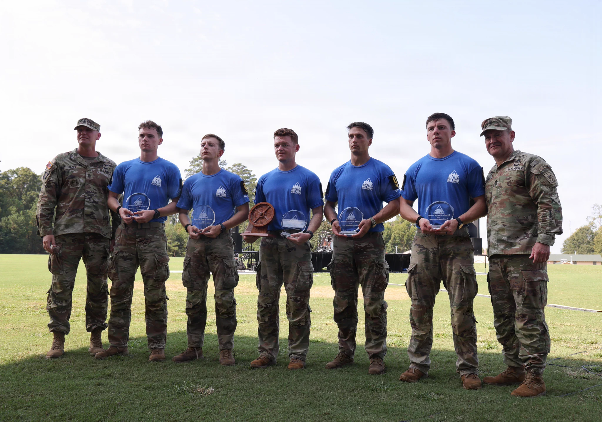 Seven men stand on grass under a cloudy sky. Two men in full Army camouflage uniforms flank five men in blue \"BEST SQUAD COMPETITION\" t-shirts and camo pants. The five hold clear \"Granite Charge Award\" plaques; one also holds a wooden barbell weight award. Names Paske & Stewart are visible.