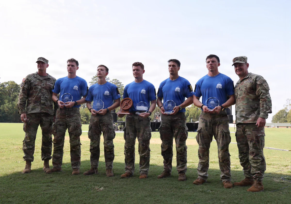 Seven men stand on grass under a cloudy sky. Two men in full Army camouflage uniforms flank five men in blue \"BEST SQUAD COMPETITION\" t-shirts and camo pants. The five hold clear \"Granite Charge Award\" plaques; one also holds a wooden barbell weight award. Names Paske & Stewart are visible.
