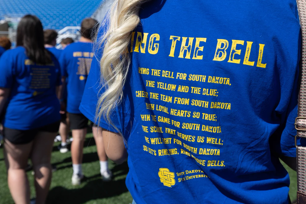 A group of people stand on a grassy field, all wearing blue South Dakota State University t-shirts. The shirt in the foreground displays yellow text with a chant about ringing a bell for South Dakota, mentioning "The Yellow and The Blue" and South Dakota State University.
