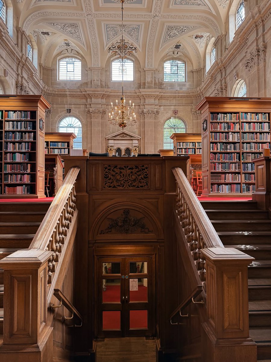 View up a grand wooden staircase into Lincoln College Library, with rows of bookshelves, a chandelier and an ornate vaulted ceiling above.