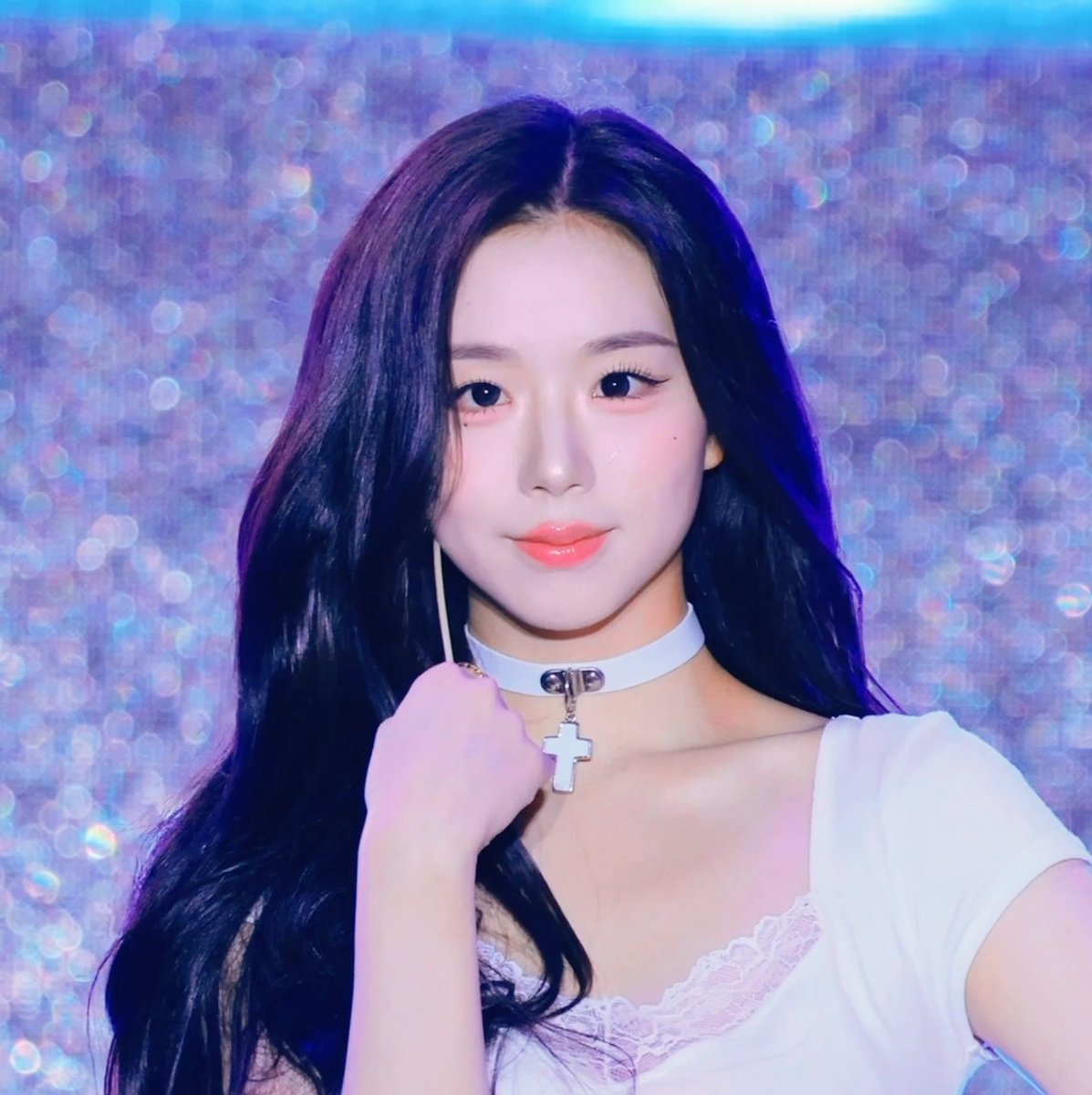 A portrait of a young woman with long dark hair wearing a white lace top and a white choker with a cross pendant, set against a sparkly purple background.
