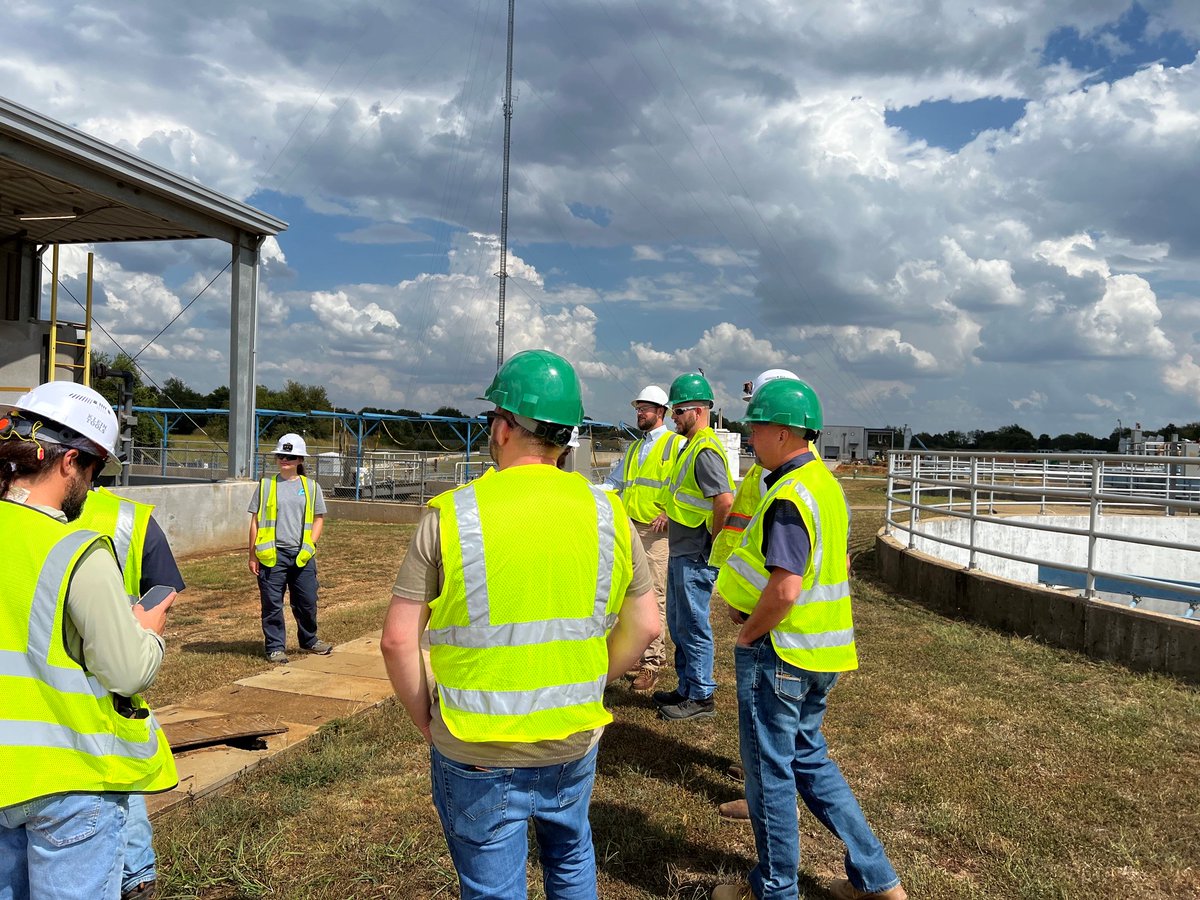 Group of construction workers wearing green and white helmets and yellow safety vests standing near a water treatment facility under a cloudy sky.