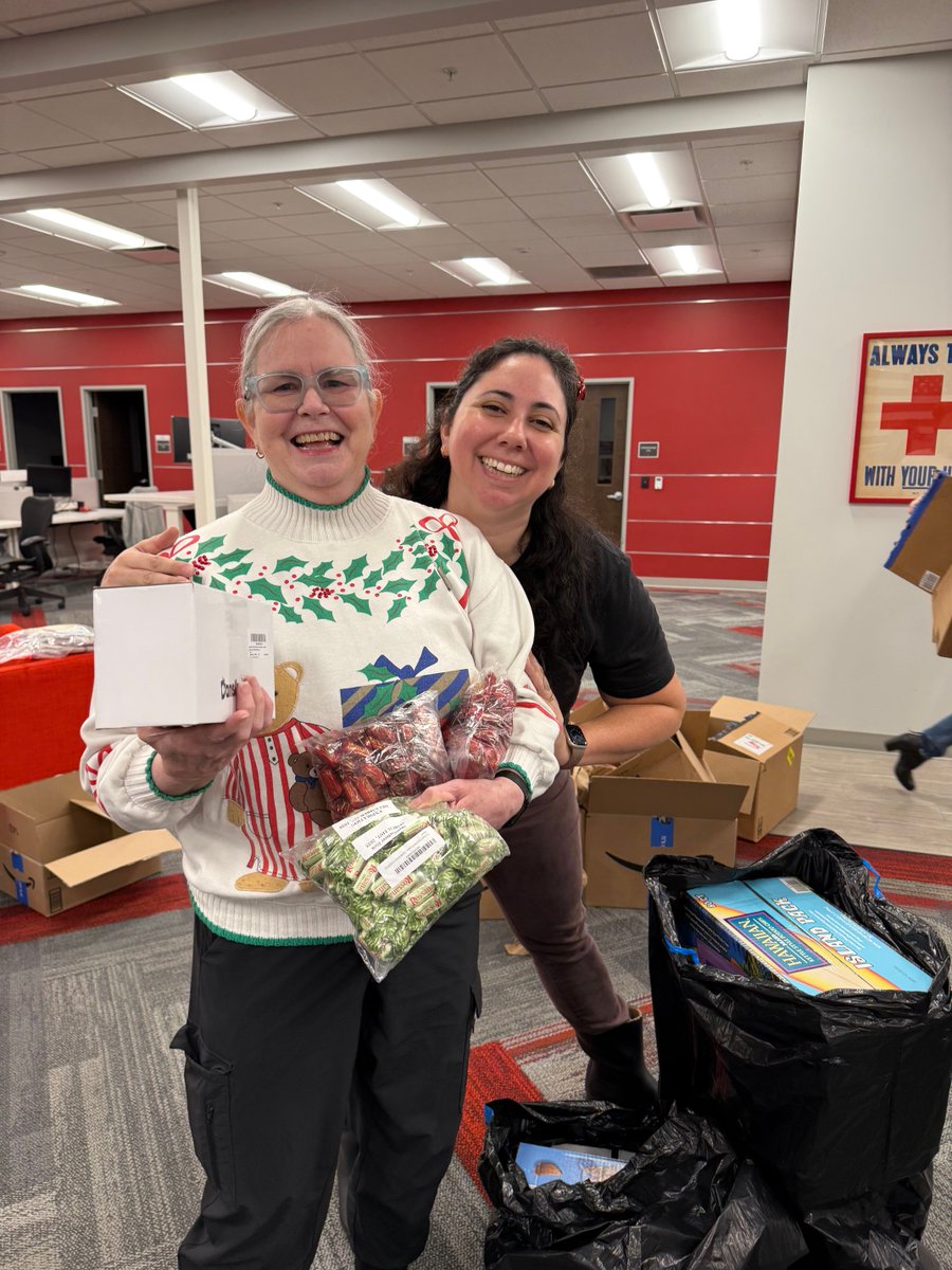 Two people in a room are smiling while holding bags of red grapes and what appears to be other food items. There are several cardboard boxes and a large black bag nearby. A first aid sign with a red cross and "Advent's Ministry" is visible on the wall in the background.