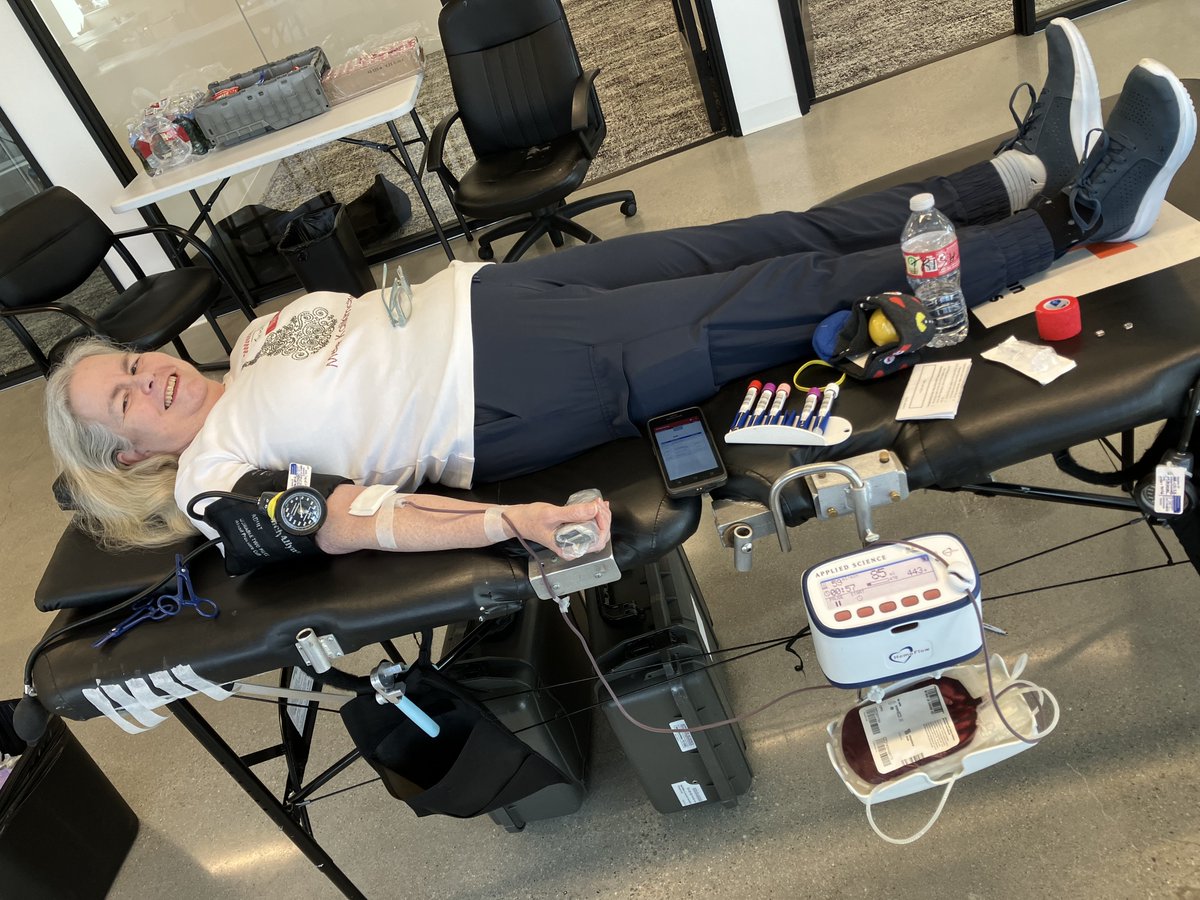 A person is lying on a blood donation chair with a blood pressure cuff on the arm. A blood donation bag and medical equipment are visible. There is a bottle of Smartwater and a can of Dr Pepper on a nearby table.