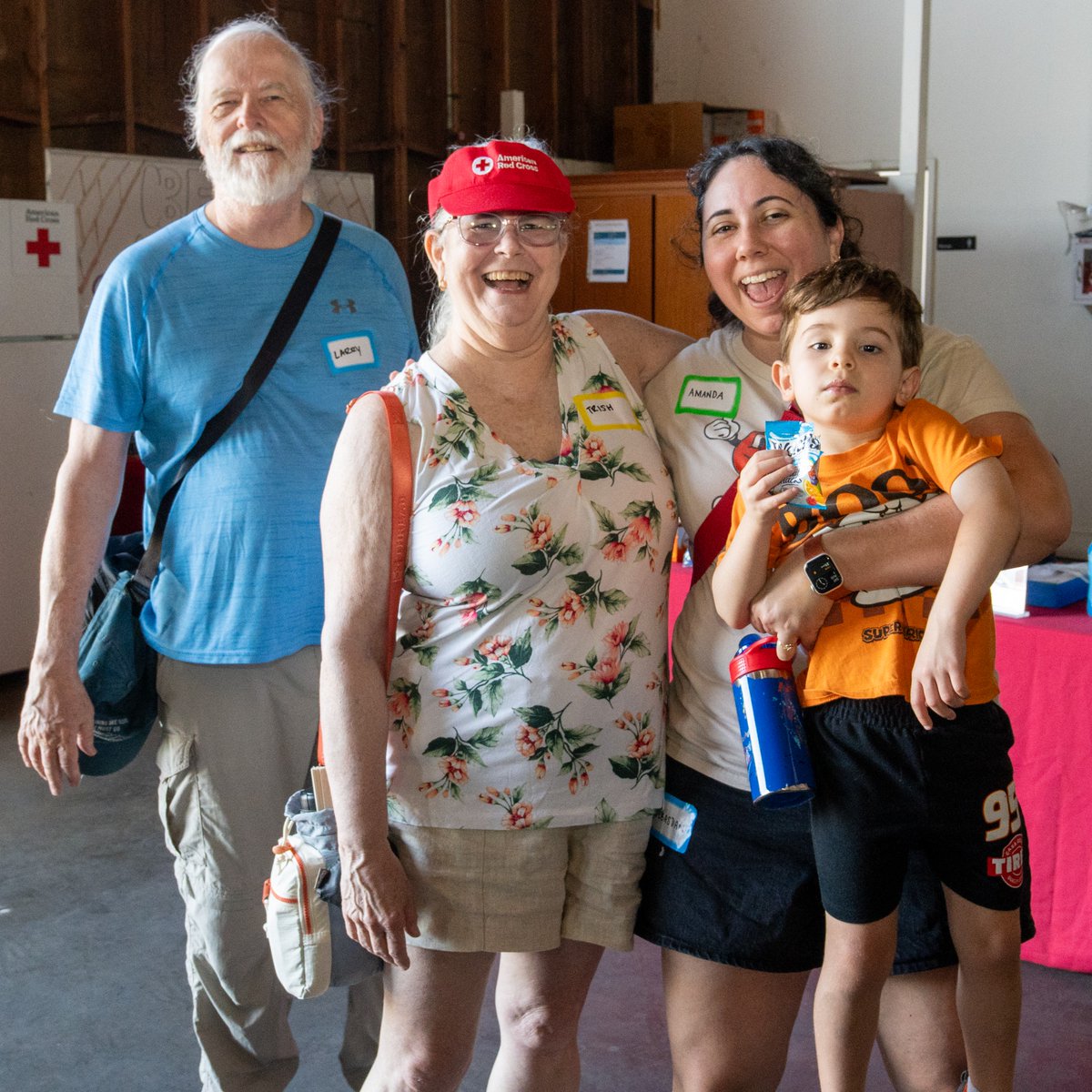 A group of four people smiling indoors. One person is holding a child. They are standing near a table with a red American Red Cross tablecloth. Two individuals are wearing name tags: "Larry" and "Karen.