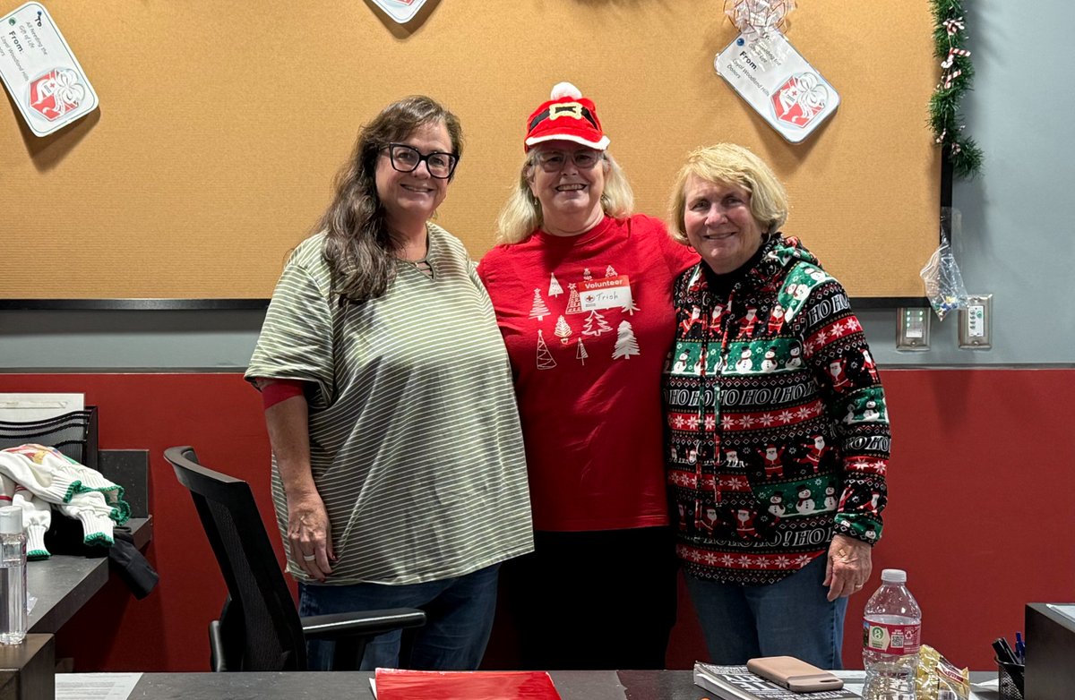 Three people stand together smiling in an office setting, featuring festive decorations and attire including Christmas-themed shirts and a Santa hat. They are surrounded by a workspace with various items like a water bottle and papers.