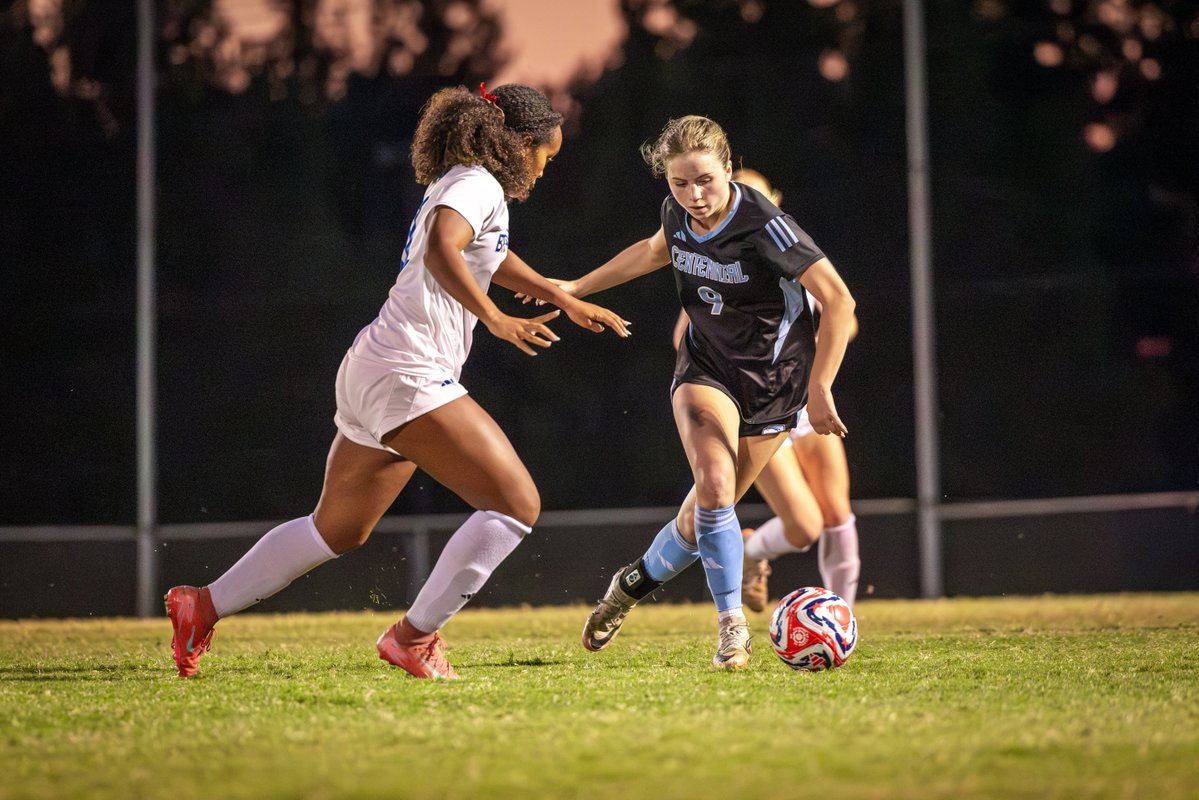 Two soccer players on a field at night, one in a light jersey and the other in a dark jersey, engaged in a close on-the-ball duel.