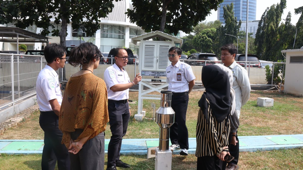 Tiga petugas pria BMKG berkemeja putih menjelaskan instrumen meteorologi kepada empat mahasiswa magang yang mengenakan pakaian berwarna cokelat, garis hitam putih, abu-abu, dan putih (kiri ke kanan). Keempatnya berdiri menghadap petugas dan bersama petugas mereka mengelilingi alat penakar hujan. Di belakang mereka terdapat alat sangkar meteorologi, pepohonan, lokasi parkir mobil, dan gedung kantor BMKG.