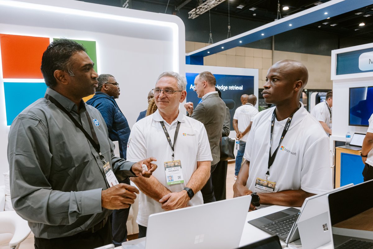 Four men engage in discussion at a Microsoft exhibition booth. Two men wear white Microsoft polo shirts, while one in a gray shirt gestures animatedly to the group. Laptops are visible on the table.