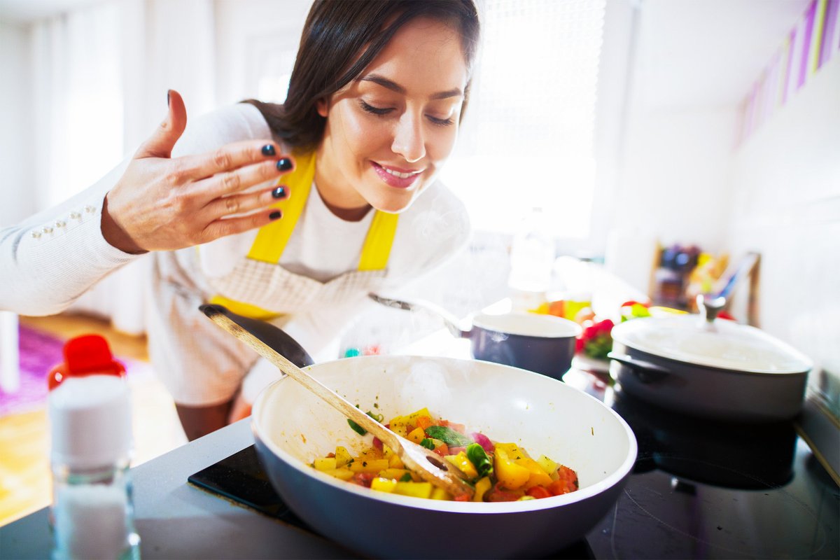 A person cooking in a brightly lit kitchen, wearing a yellow apron. They are leaning over a pan on the stove, filled with colorful vegetables, appearing to enjoy the aroma.