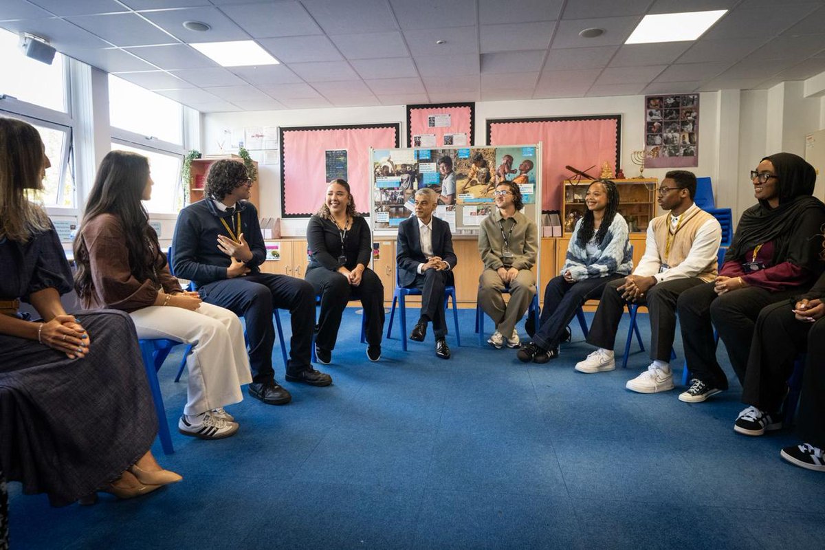 Mayor of London Sadiq Khan sits with secondary school students in a classroom discussion. Actress Nkechi Simms, wearing a blue sweater, is seated among the students. The group sits in a circle in a bright classroom.
