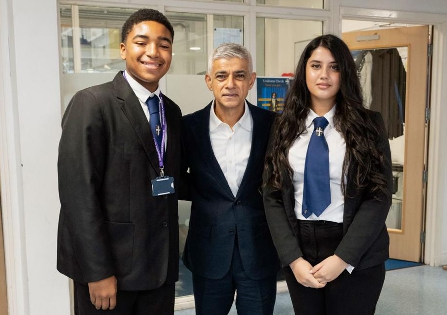 Mayor of London Sadiq Khan poses for a photograph between two secondary school students, one male student and one female student, in a school corridor. All three are facing the camera.