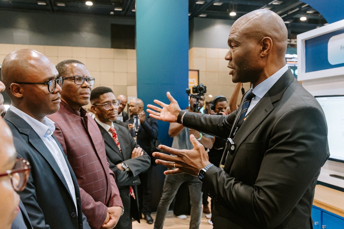 A group of professionals in business attire attentively listens to a speaker at a conference. The speaker, a man in a suit, gestures expressively while explaining something to three men. One wears glasses and another has a patterned shirt.