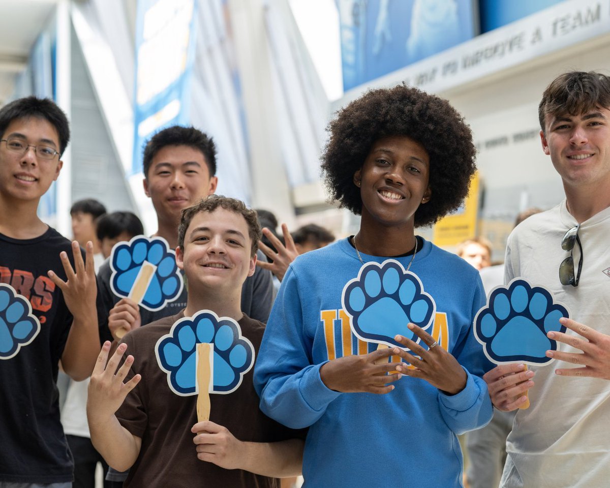 Five students stand together smiling and holding blue UCLA paw signs inside Pauley Pavilion.
