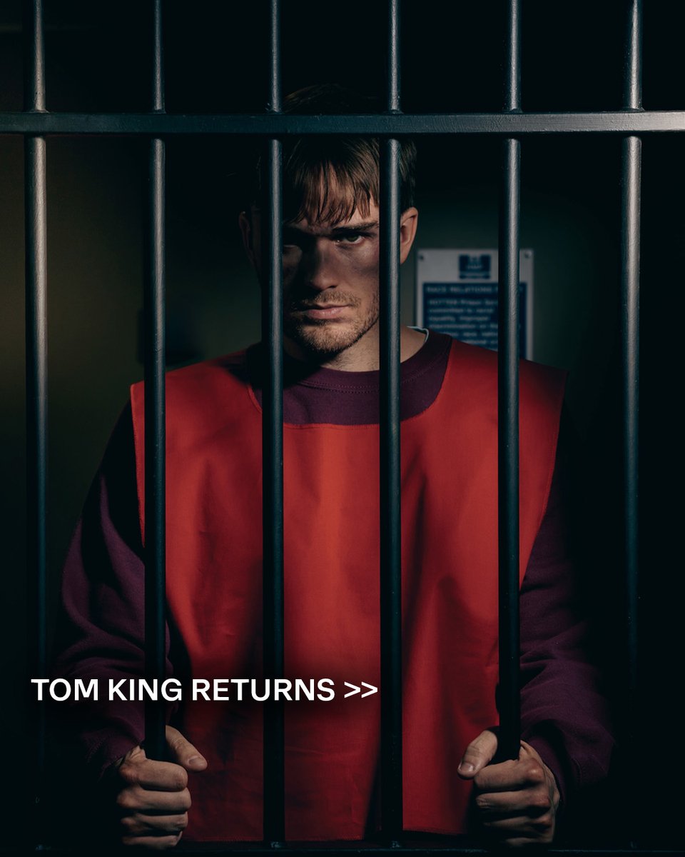 Tom King standing behind prison bars on a prison jumper and prison red bib.\n\nHe's looking straight down the camera with a menacing facial expression.