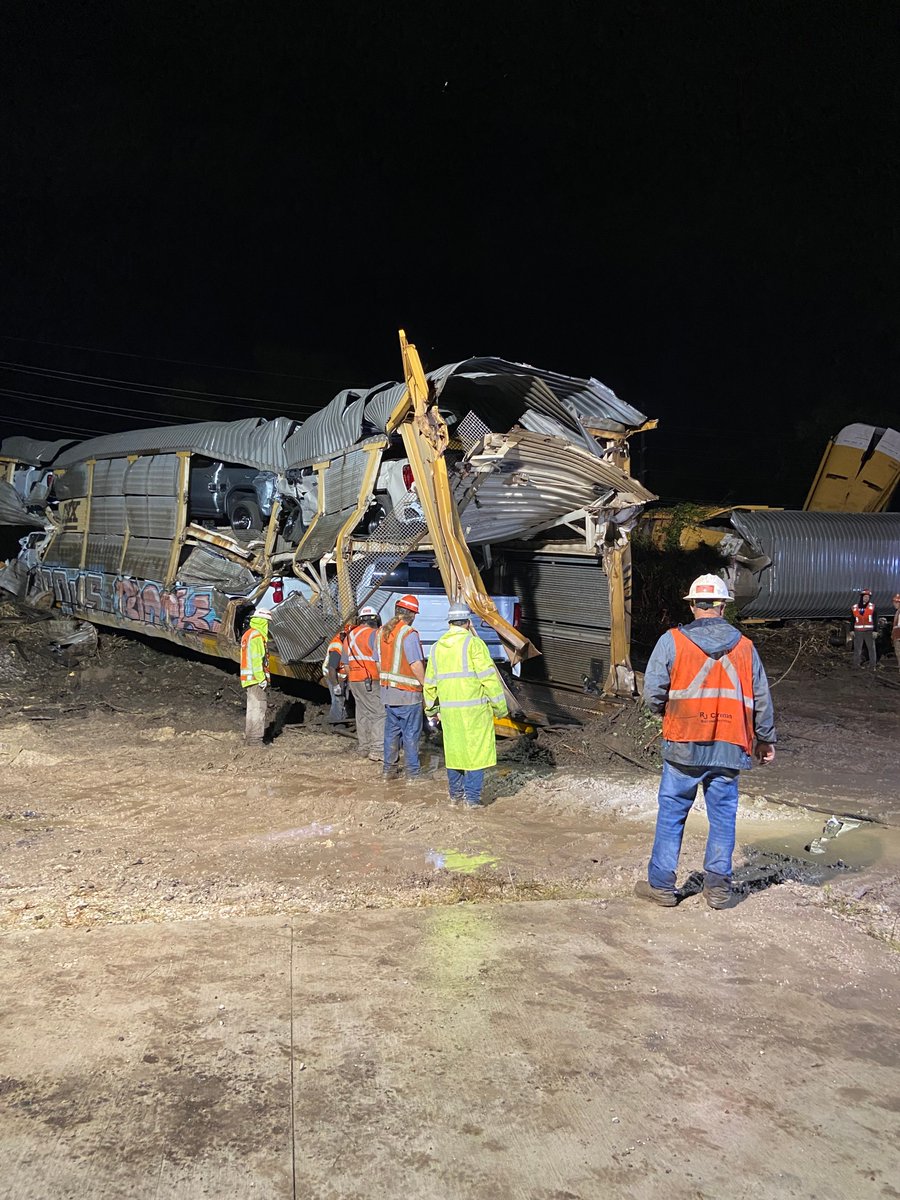 Workers in safety gear inspect a severely damaged train car at night on a dirt site after a derailment.