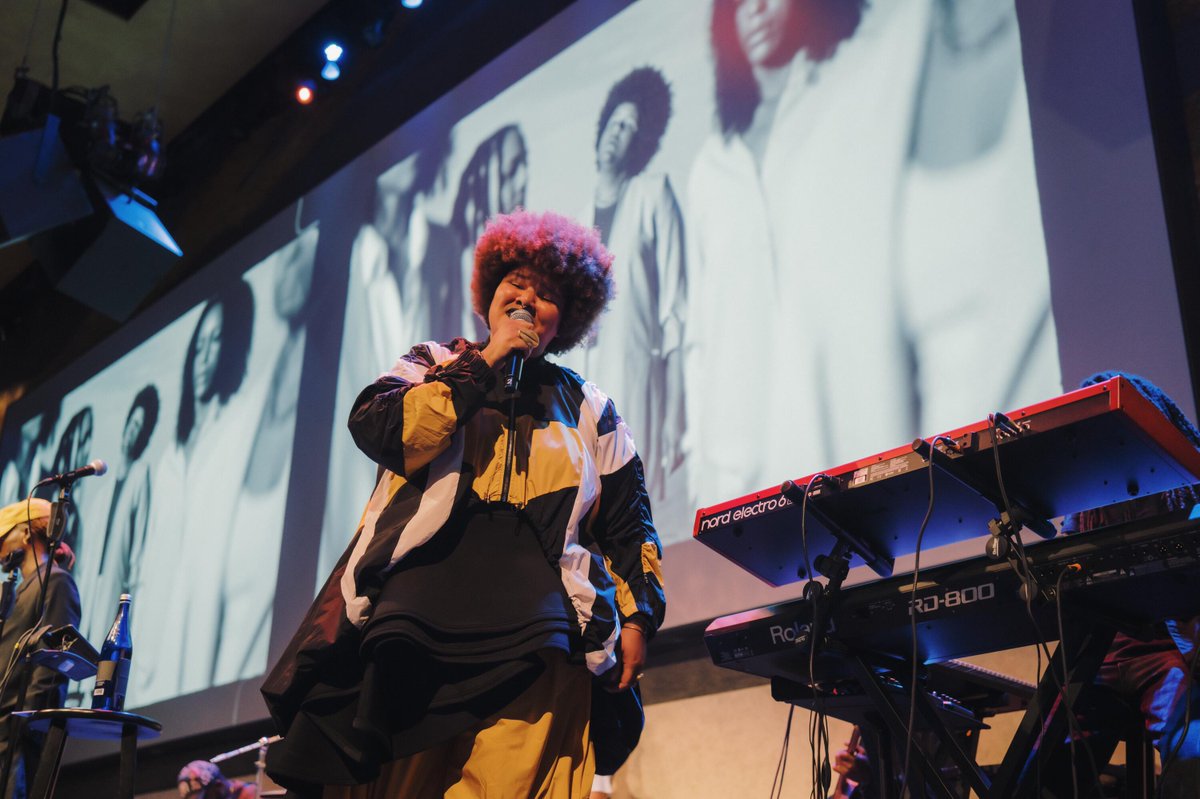A performer sings on stage, holding a microphone. A large screen behind displays a grayscale image of the performer. A keyboard is visible on the right side of the stage.