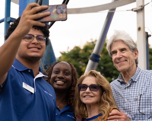 A student holds up a phone to take a selfie with another student, a woman in sunglasses and an older man in a blue checkered shirt near a carnival ride.