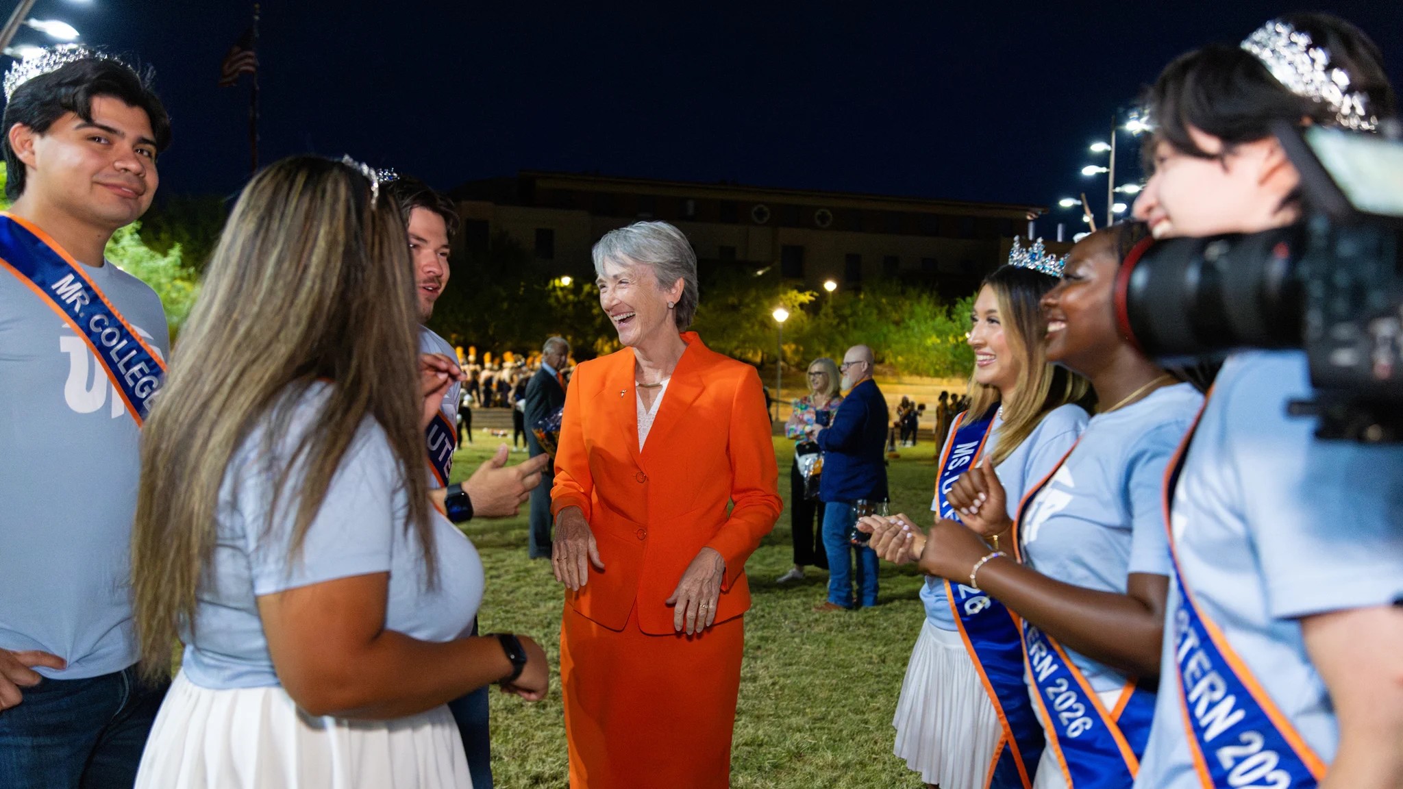 The 2026 UTEP Homecoming Court is gathered around university president Heather Wilson in an orange suit, smiling and conversing outdoors on the UTEP campus.