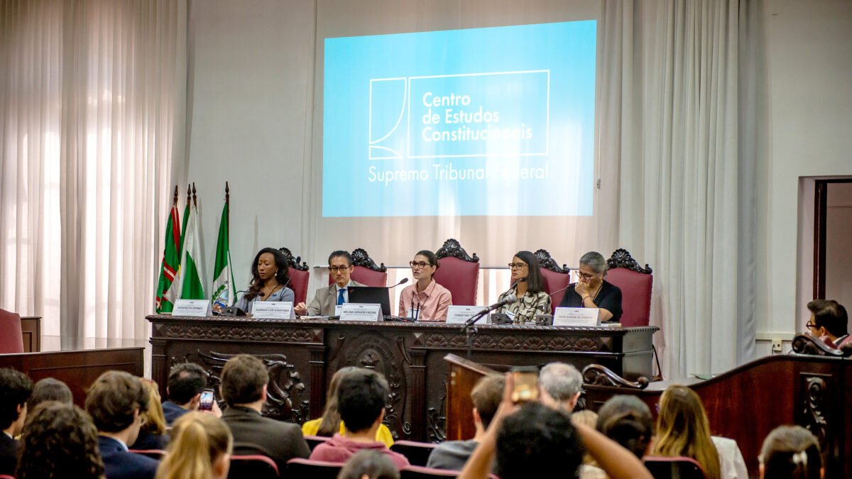 Fotografia horizontal de uma audiência acadêmica do Centro de Estudos Constitucionais do STF. Há uma bancada com representantes da iniciativa e pessoas acompanhando a conversa. Fim da descrição.