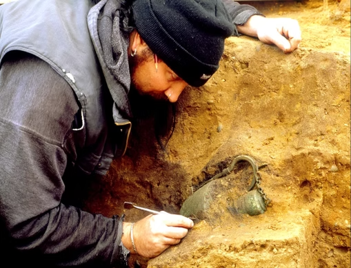 AngloSaxonPast's tweet photo. A road scheme uncovered England’s earliest dated Anglo-Saxon princely burial
In 2003, during the early Anglo-Saxon conversion period, archaeologists checking land for a proposed road-widening scheme at Prittlewell in Southend-on-Sea, Essex, uncovered something they had no reason to expect: an almost undisturbed timber burial chamber dating to about AD 580–605. 
On the first day, a large square pit appeared. Then came the moment that changed the excavation. A copper-alloy bowl emerged still hanging from an iron hook where a wooden chamber wall had once stood.
The wood itself had mostly vanished. Its stains had not.
Those stains allowed archaeologists to trace a room roughly four metres square. A coffin had stood inside it, surrounded by a sword, shield, drinking vessels, gaming pieces, a folding stool and objects that had travelled extraordinary distances. Some came from the eastern Mediterranean. Garnets used on one object probably originated in India or Sri Lanka. v
Yet the person at the centre of all this wealth had almost disappeared. Acidic soil destroyed nearly the entire skeleton, leaving only tiny fragments of tooth enamel. Weapons and dress fittings suggest the burial was probably male, but even that conclusion rests on objects rather than surviving bones. His name is unknown. 
Then two scraps of gold complicated the story further.
Near the head of the coffin lay two thin gold crosses, probably placed over the dead person's eyes. They point towards Christianity, while the lavish furnished burial belonged to older funerary traditions. The burial's date overlaps the years around Augustine's Roman mission of AD 597, showing that Christian ideas were already moving through elite networks in ways a simple conversion timeline cannot capture. 
One of the quietest finds may be the most human.
A wooden lyre had decayed almost completely into a dark shape in the soil. Archaeologists lifted the entire block and scanned it. The outline revealed a maple instrument with ash tuning pegs and garnet fittings.
More importantly, it had broken during its working life.
Someone had carefully repaired it with iron, gilded copper alloy and silver, then kept using it before it finally went into the grave.