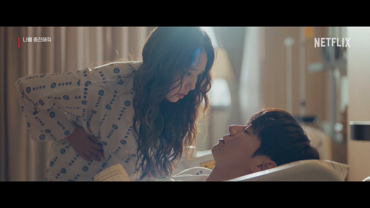 A woman in a hospital gown leans intimately over a man lying in a hospital bed, gazing into each other's eyes in a soft-lit room, with the "Netflix" logo and "나를 충전해줘" (Charge Me Up) text visible on screen—capturing the romantic spark teased in the preview between the "electric ability user" Naboba and her "human rubber glove" Baekho.