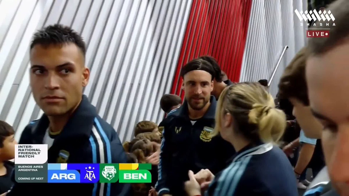 Two Argentine national team players stand in a corridor before an international friendly match, with on-screen graphics displaying "ARG vs BEN" for an upcoming match in Buenos Aires, and "SHASHA LIVE" branded in the top right corner, as referenced in the post about Messi's pre-match walk onto the pitch.