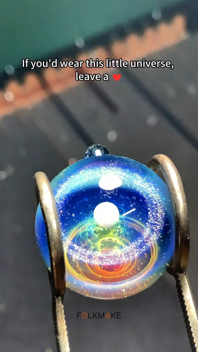 A glass sphere containing a miniature, colorful galaxy is held by tweezers, with overlay text reading 'If you'd wear this little universe, leave a ❤️' and 'FOLKMAKE'.