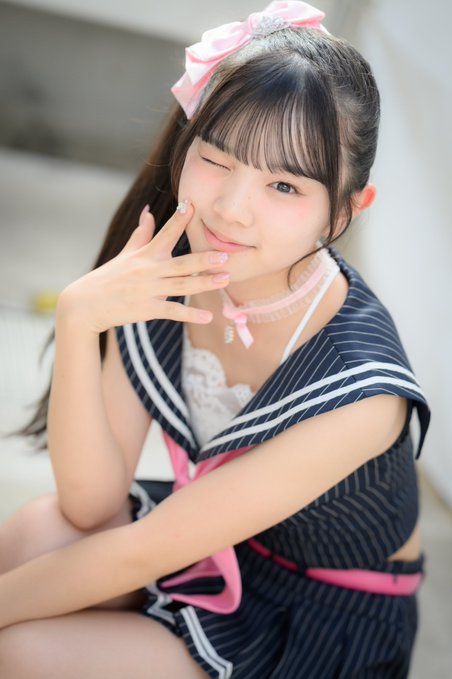 A young woman with a sailor uniform-style outfit and a pink bow headband winks at the camera while holding her fingers near her cheek.