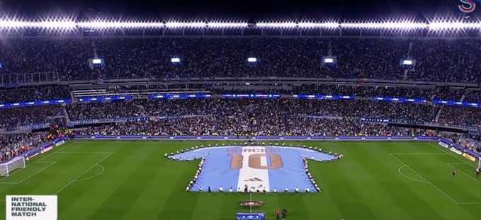 A packed soccer stadium at night hosts an "INTERNATIONAL FRIENDLY MATCH" between Argentina and Chile, as indicated by the giant jersey-shaped banner on the pitch displaying "FIFA" with the two national flags. The "Blessed Mantle" mentioned in the post likely refers to the massive ceremonial jersey banner draped across the field, which resembles a holy cloak draped over the stadium.