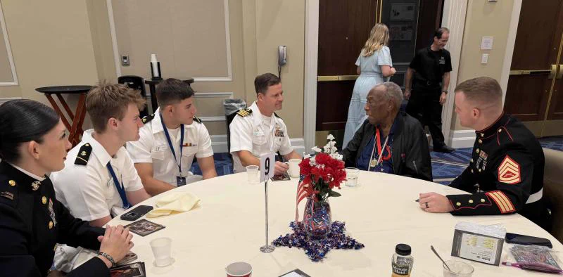 A group of people in uniform sit around a round table decorated with patriotic-themed centerpieces. They appear to be engaged in conversation. A person wearing a medal sits with them. In the background, a person in civilian clothing walks by.