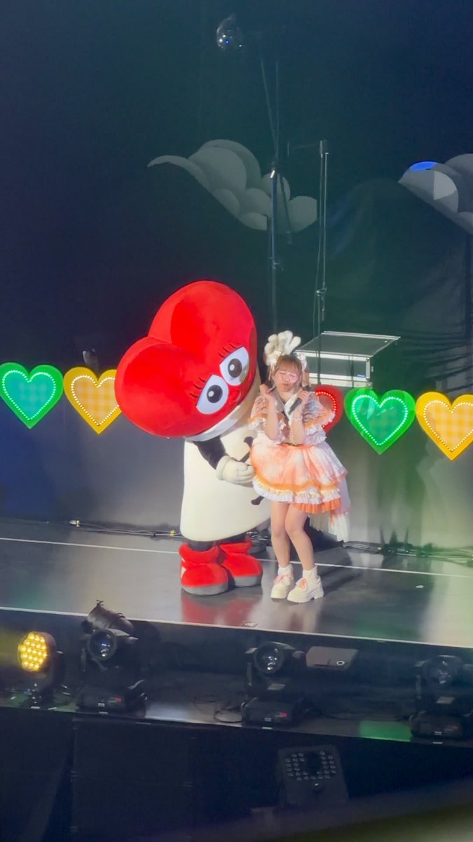 A young woman in a frilly orange and white outfit next to a large red mascot on a stage decorated with colorful heart lights.