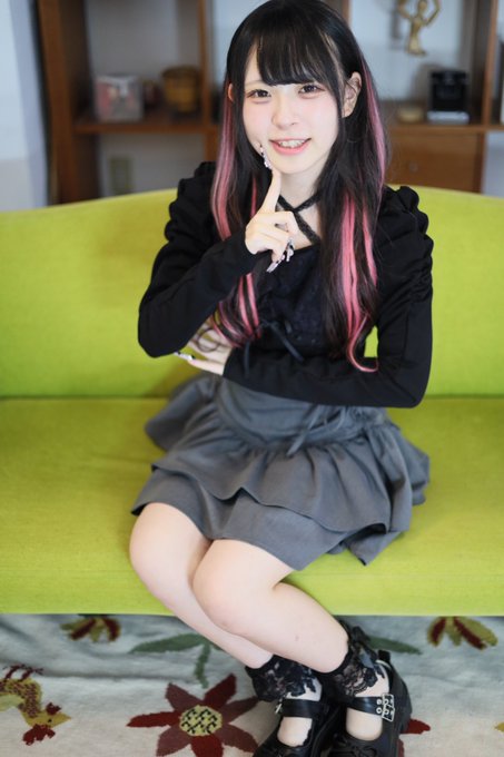 A young woman with black hair featuring pink highlights sits on a green sofa, wearing a black top and grey skirt, making a fist gesture.