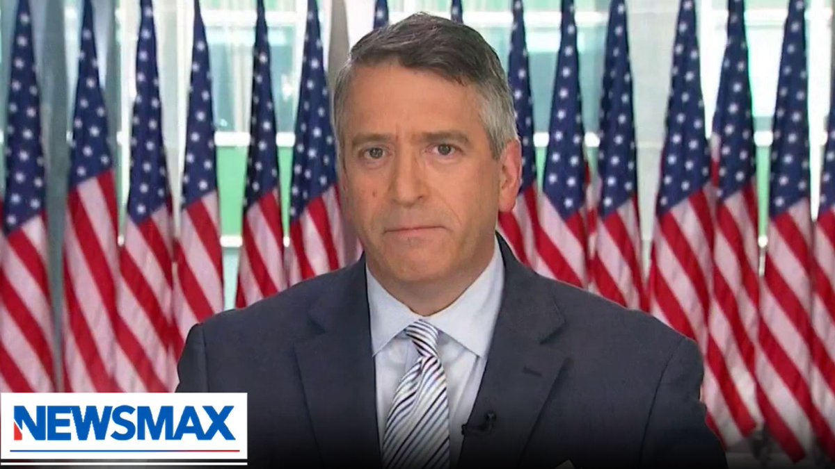 A gray-haired man in a dark suit and striped tie is shown on a Newsmax broadcast with American flags displayed in the background, appearing as a guest commentator on the network's coverage of the rumored pneumonic plague outbreak story.