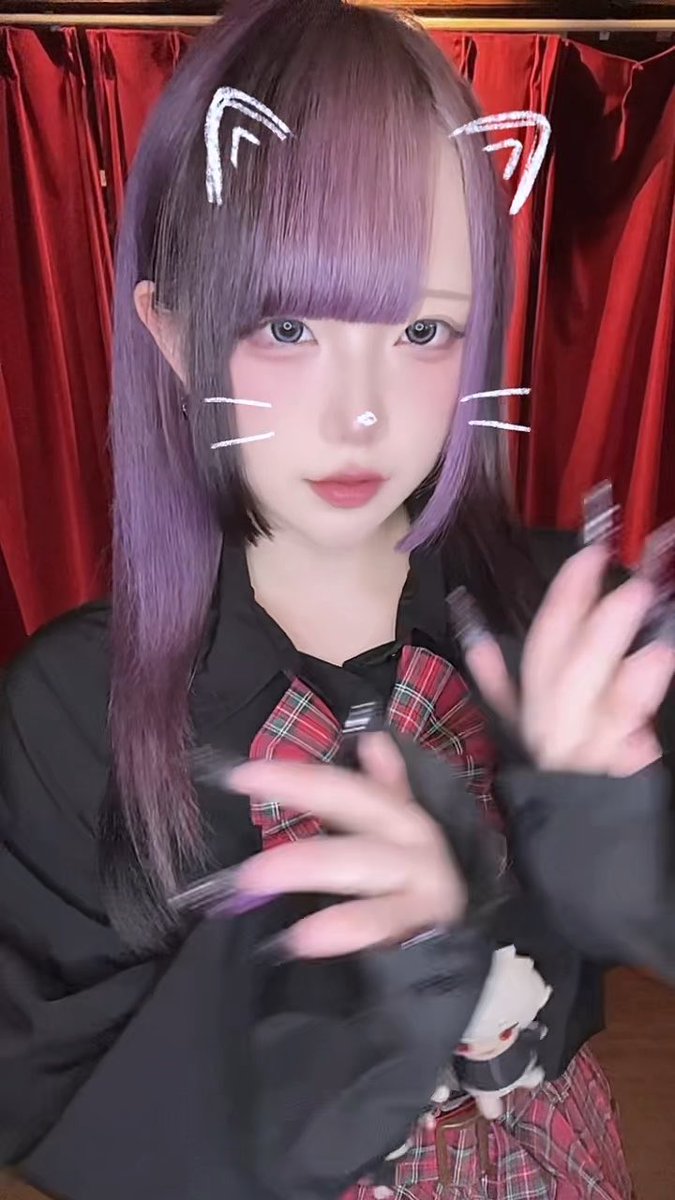 A young woman with purple hair and bangs, wearing cat ear and whisker face filters, a black top, and a red plaid tie, poses in front of a red curtain.
