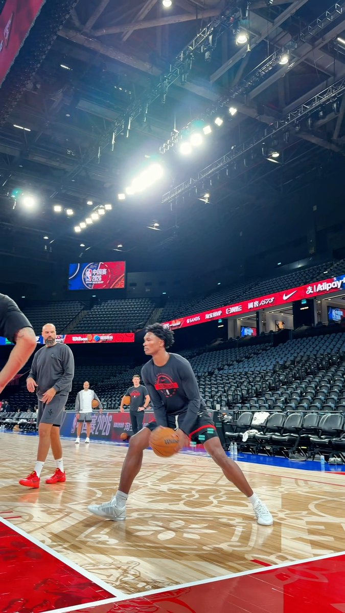 A basketball player practices a floater or jump shot on a uniquely patterned court during a pre-game warmup at what appears to be the NBA China Games 2026, as displayed on the arena's center-hung scoreboard.