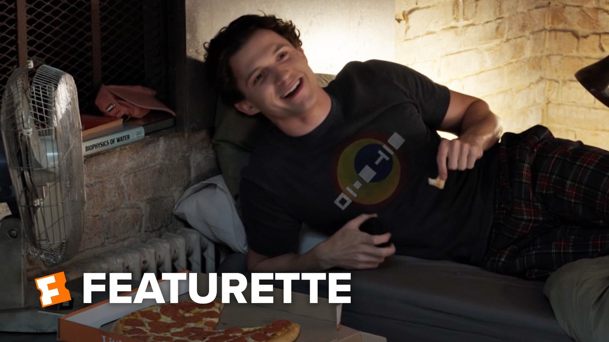 A young man lies on a bed in a brick-walled room next to a pizza box and a book titled 'BIOPHYSICS OF WATER', with 'F FEATURETTE' text on screen.